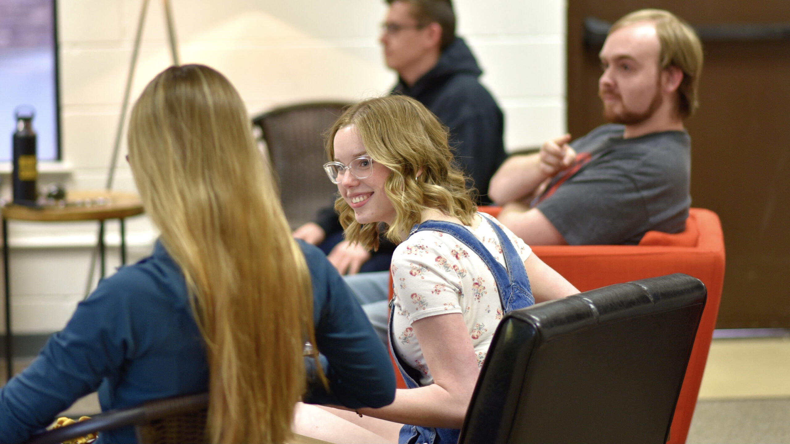 A smiling young woman with glasses sitting in a group of friends leaning forward to listen to the person sitting next to her.