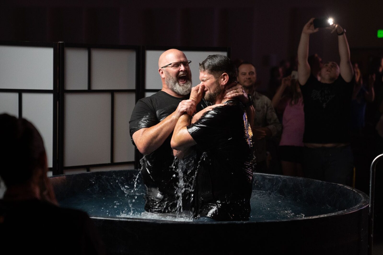 A man coming out of the water holding his nose while being baptized by a tall smiling man with a gray beard